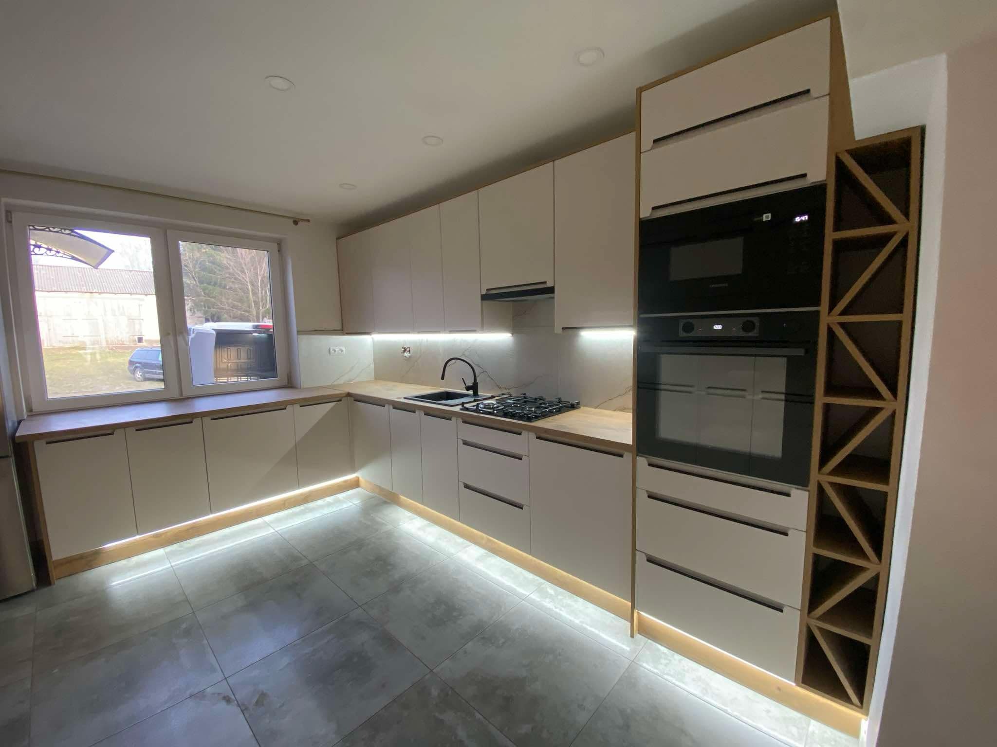 Jasna kuchnia z oświetleniem LED pod szafkami, czarnym zlewem i płytą gazową. Uwagę przykuwa regał na wino z geometrycznym wzorem obok piekarnika.