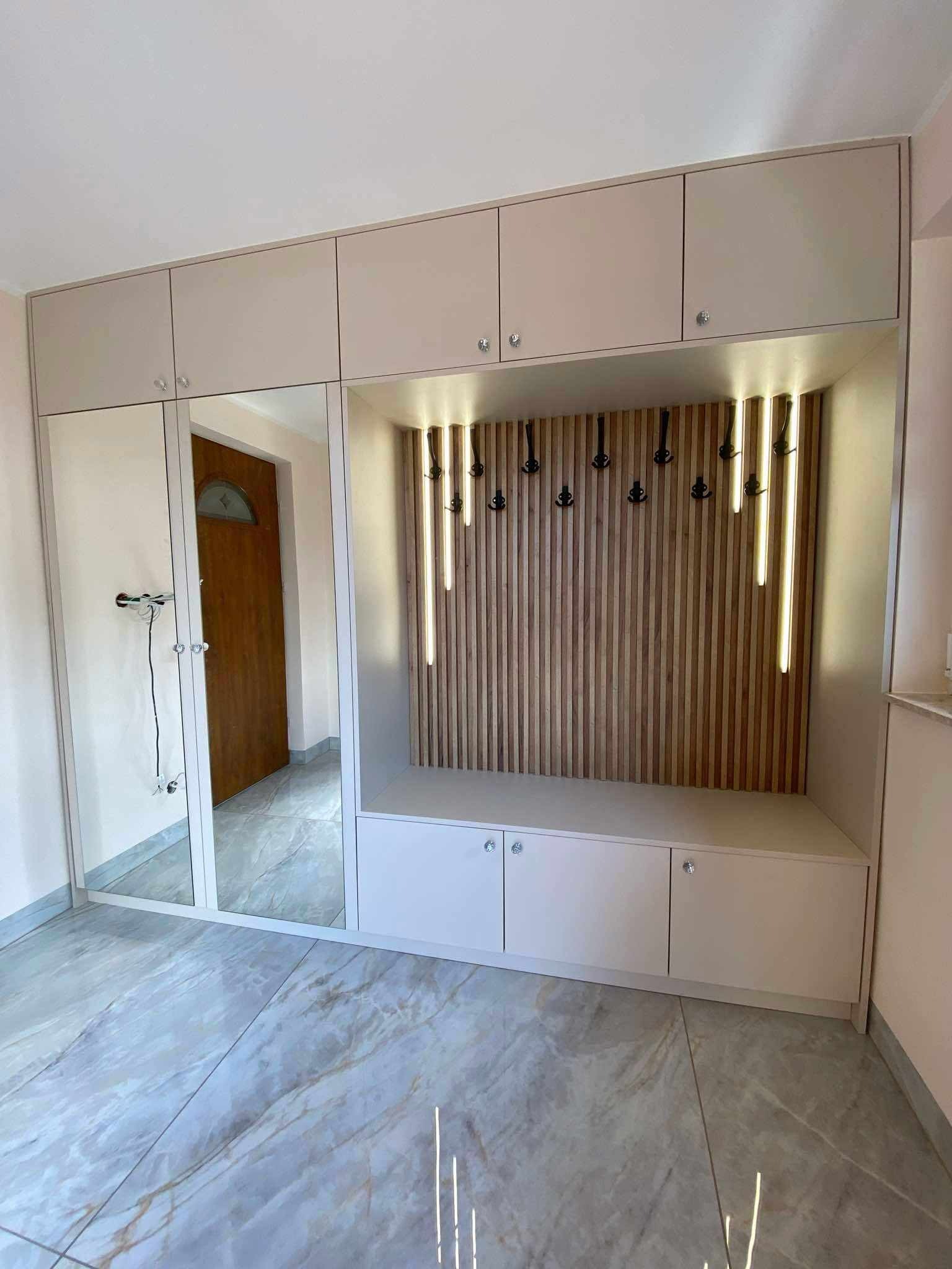 Jasna zabudowa przedpokoju z lustrami i siedziskiem z wieszakami, oświetlona pionowymi lampami. Widoczna podłoga z marmurowym wzorem i drzwi wejściowe w odbiciu.