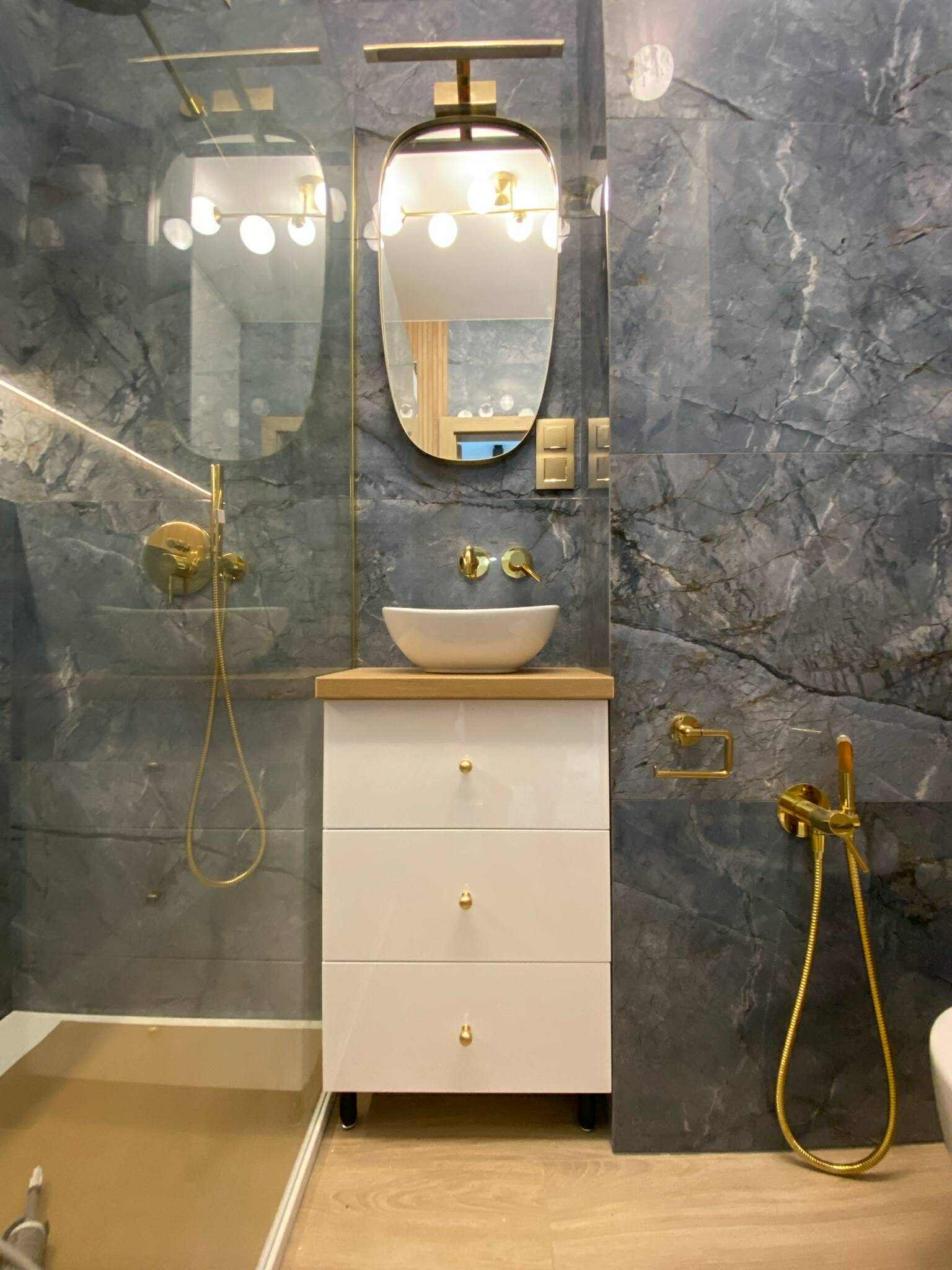 Nowoczesna łazienka z umywalką nablatową, białą szafką z trzema szufladami i złotą armaturą na tle ściany z szarego marmuru. Widoczny fragment kabiny prysznicowej.