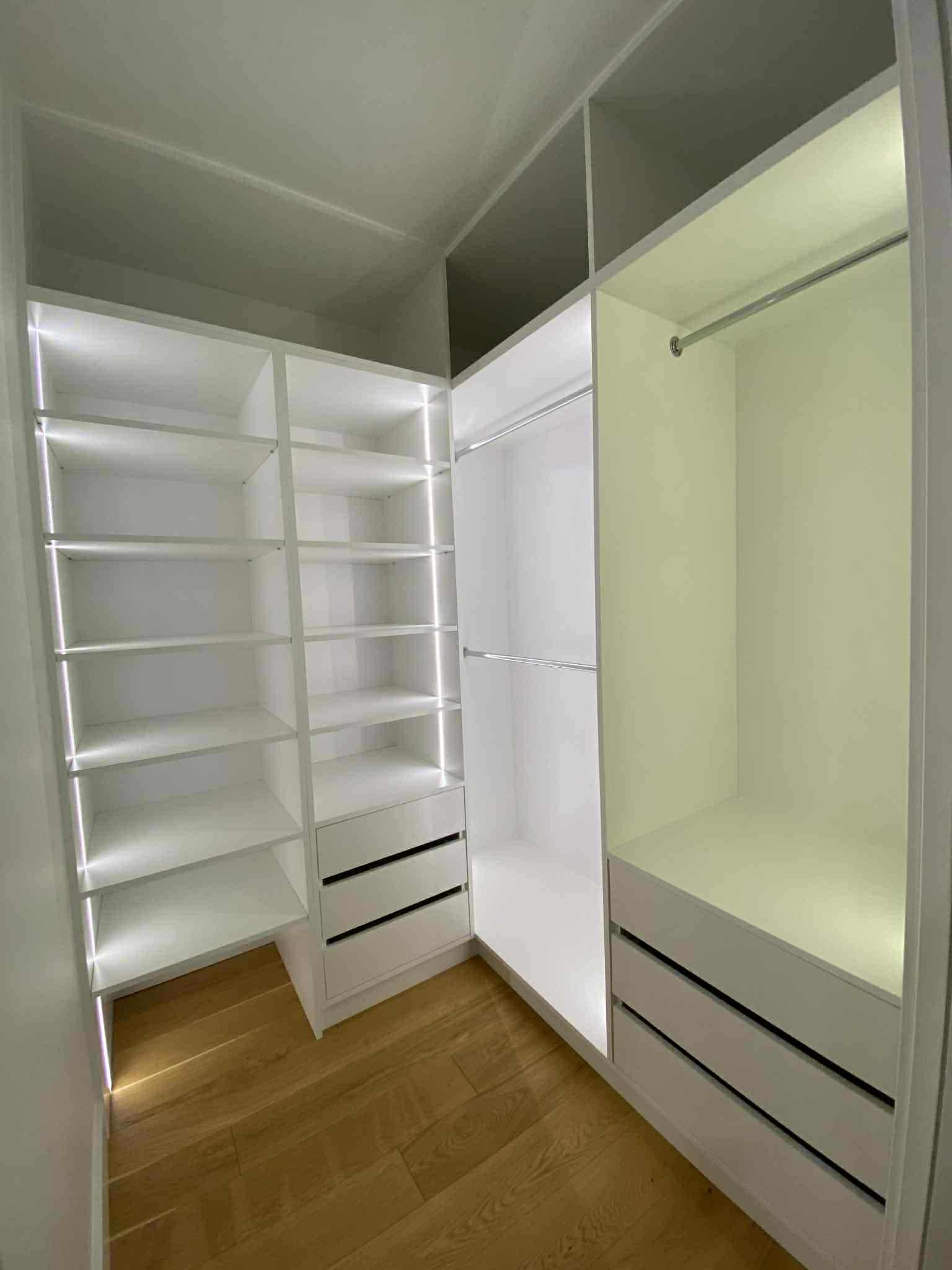 Biała szafa wnękowa z oświetleniem LED, widok z perspektywy wnętrza, drewniana podłoga. Szafa z półkami, szufladami i drążkami na ubrania.