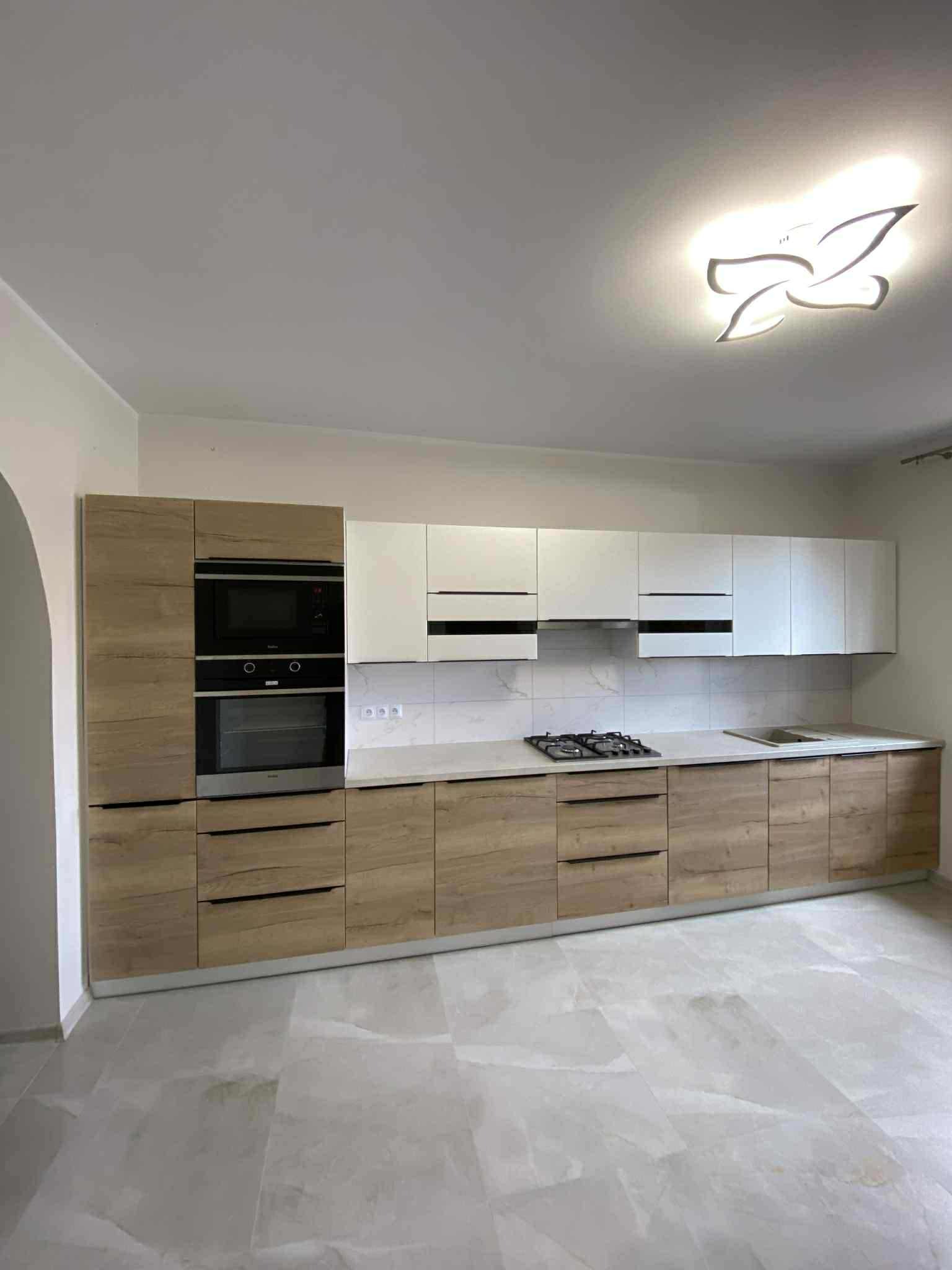 Nowoczesna kuchnia z frontami w odcieniach drewna i bieli, z wbudowanym piekarnikiem i płytą gazową, oświetlona designerską lampą sufitową.