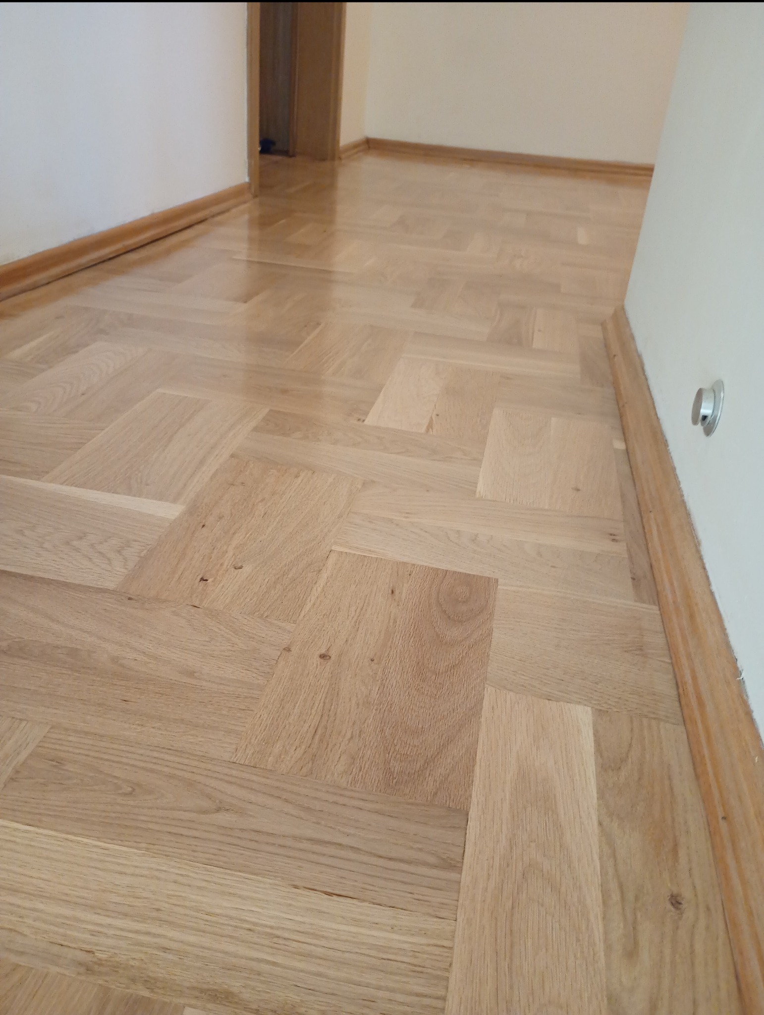 Ułożony parkiet w jodełkę francuską, wykończony drewnianymi listwami przypodłogowymi. Widoczny fragment ściany z gniazdkiem i drzwi w tle.