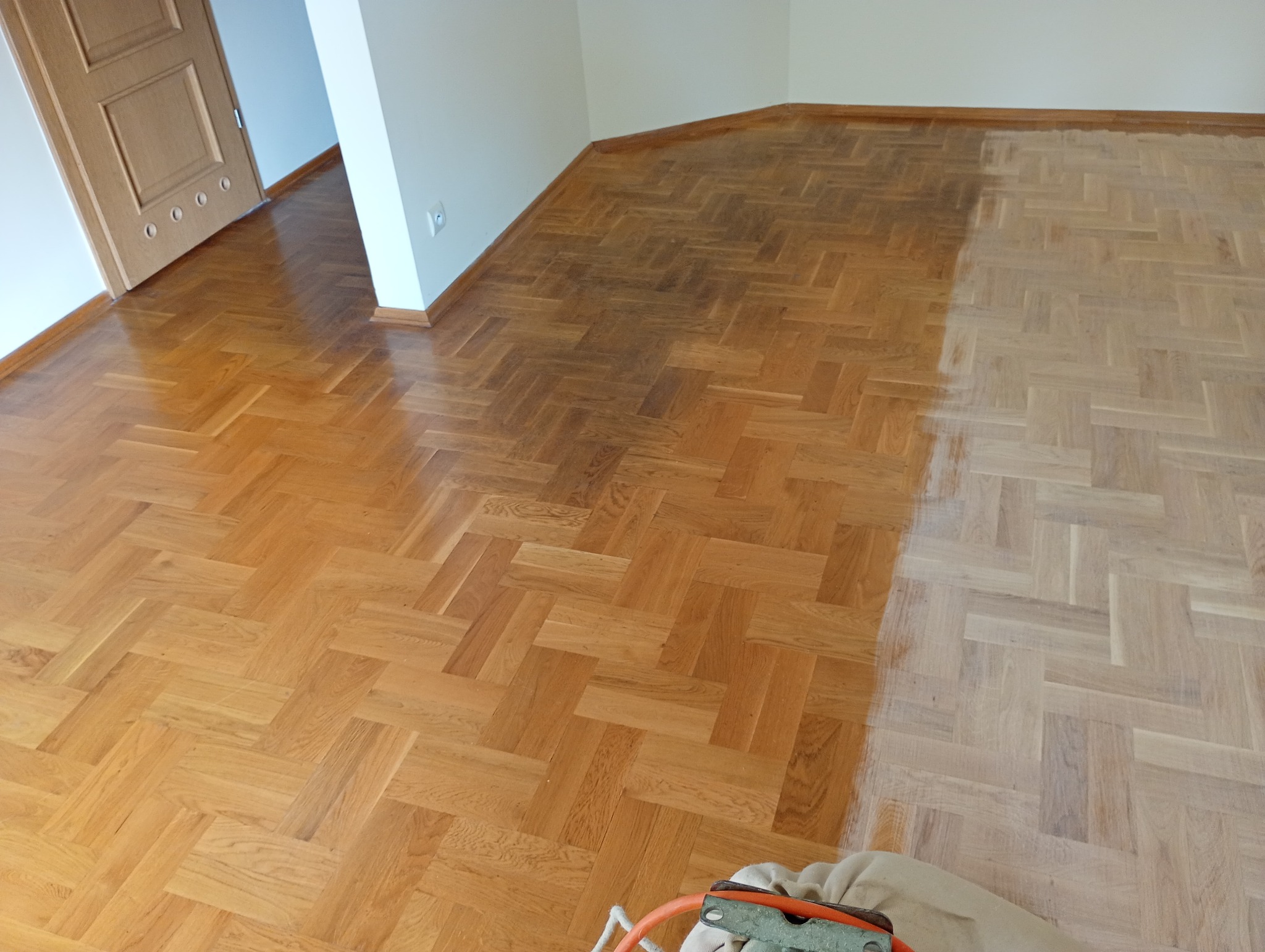 Parkiet w jodełkę częściowo odnowiony, widoczna różnica w kolorze drewna. W tle drzwi i białe ściany. Na pierwszym planie fragment cykliniarki.