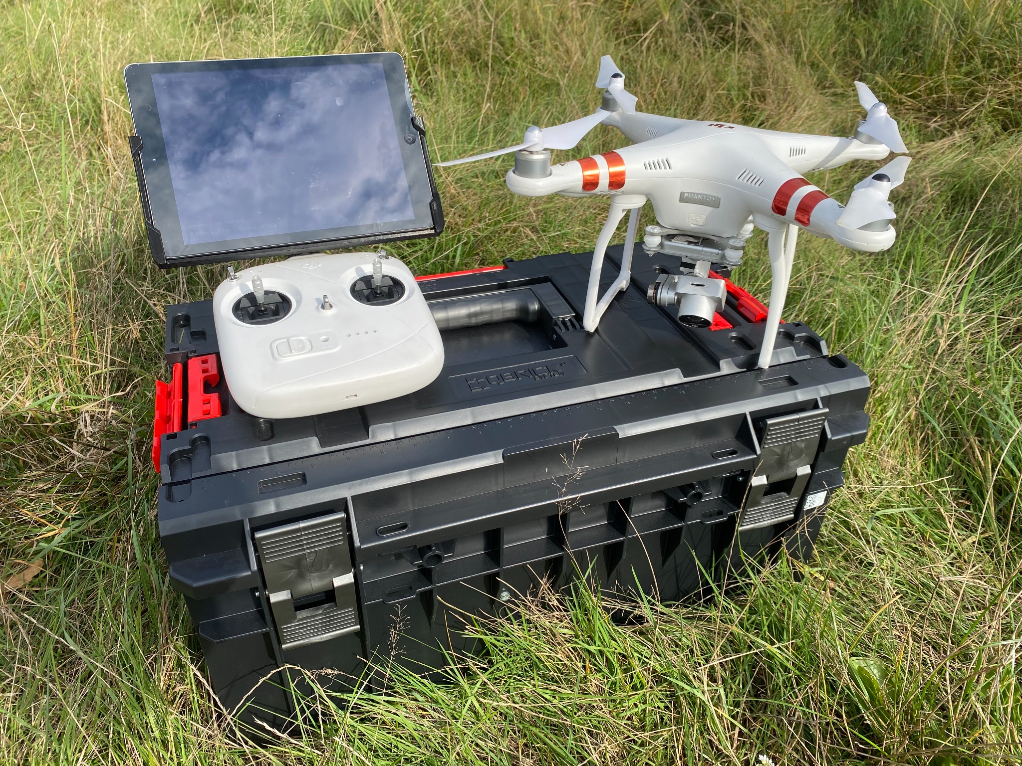 Biały dron Phantom z pomarańczowymi akcentami, kontroler i tablet leżą na czarnej skrzynce narzędziowej na tle trawy. Sprzęt gotowy do inspekcji z powietrza.