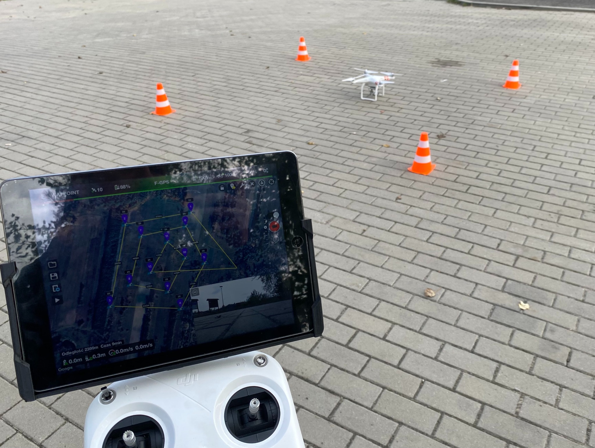 Kontroler drona z tabletem wyświetlającym mapę lotu, dron w locie między pachołkami na brukowanym placu. Widok z góry, neutralne oświetlenie.