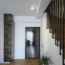 Nowoczesne wnętrze z czarną, minimalistyczną balustradą schodową i przeszklonymi drzwiami. Parkiet w jodełkę, szafka z ekspozycją szkła. Jasne, przestronne pomieszczenie.