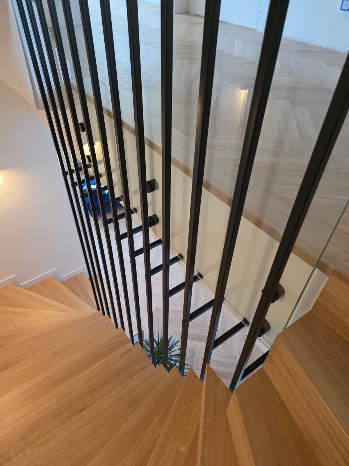 Nowoczesne schody drewniane z czarną, metalową balustradą i szklanym wypełnieniem. Widok z góry, podkreślający geometrię i minimalistyczny design.