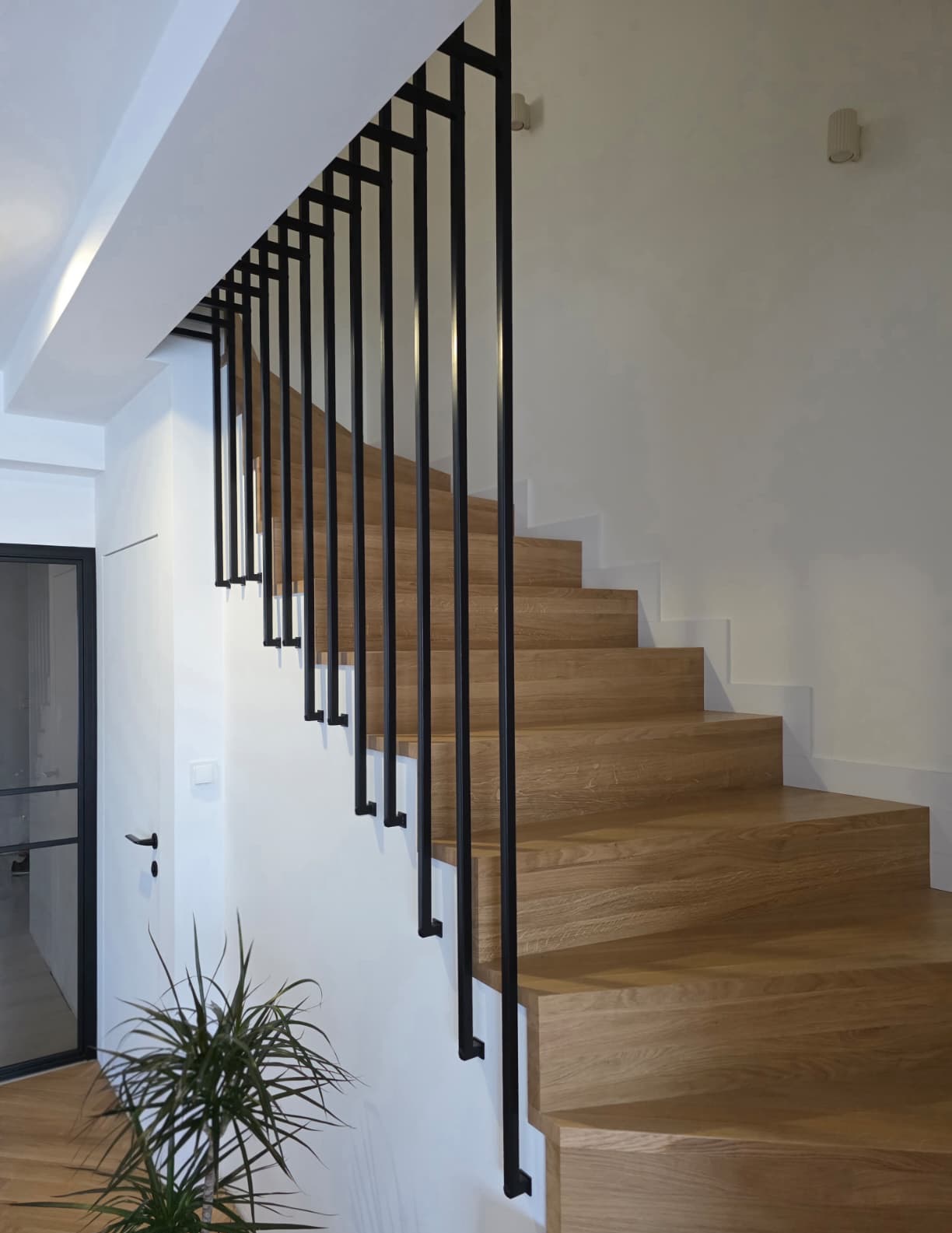 Drewniane schody z czarną, minimalistyczną balustradą. Nowoczesny design, jasne wnętrze. Ujęcie z dołu, widoczna roślina doniczkowa i fragment drzwi.