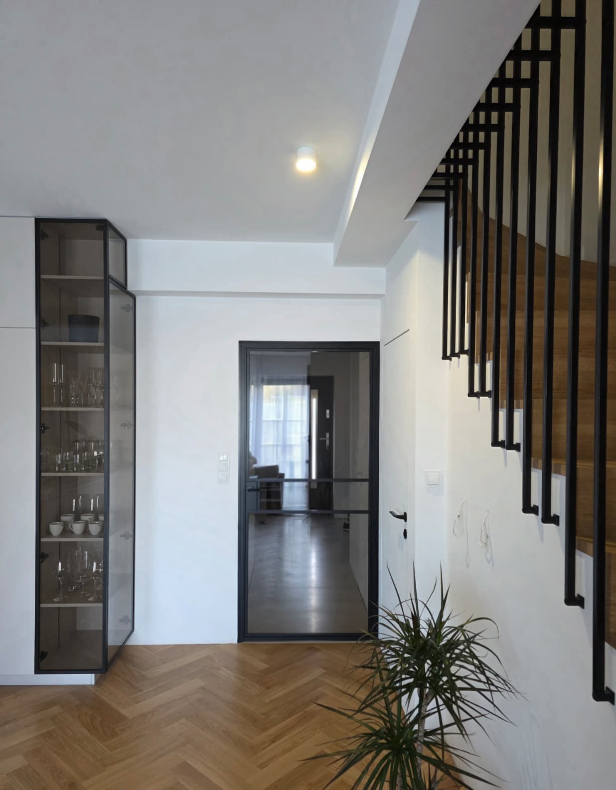 Nowoczesne wnętrze z czarną, minimalistyczną balustradą schodową i przeszklonymi drzwiami. Parkiet w jodełkę, szafka z ekspozycją szkła. Jasne, przestronne pomieszczenie.