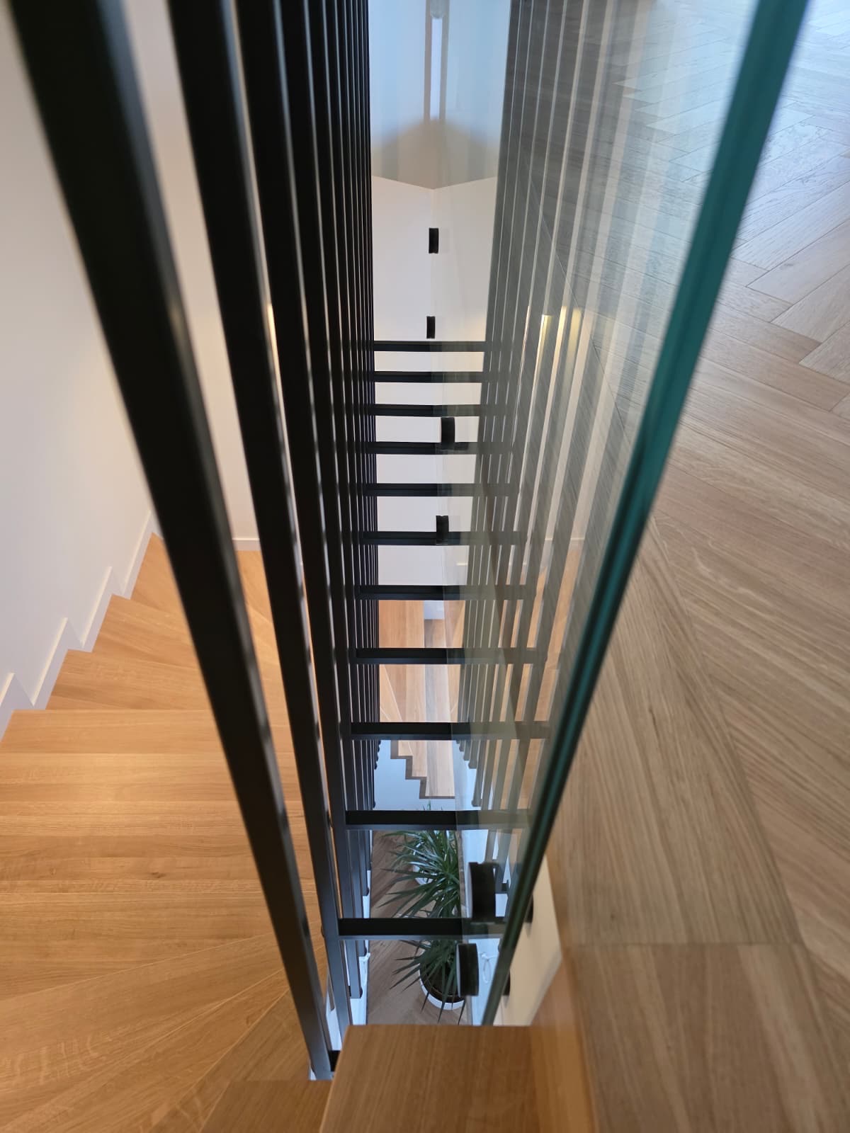 Nowoczesne schody z drewnianymi stopniami, czarną metalową balustradą i szklanym panelem, widziane z góry. Minimalistyczny design, jasne wnętrze, parkiet w jodełkę.