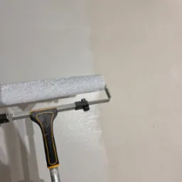 Wałek malarski z teleskopową rączką w trakcie malowania ściany na jasny odcień szarości, widoczna różnica w kolorze przed i po malowaniu.