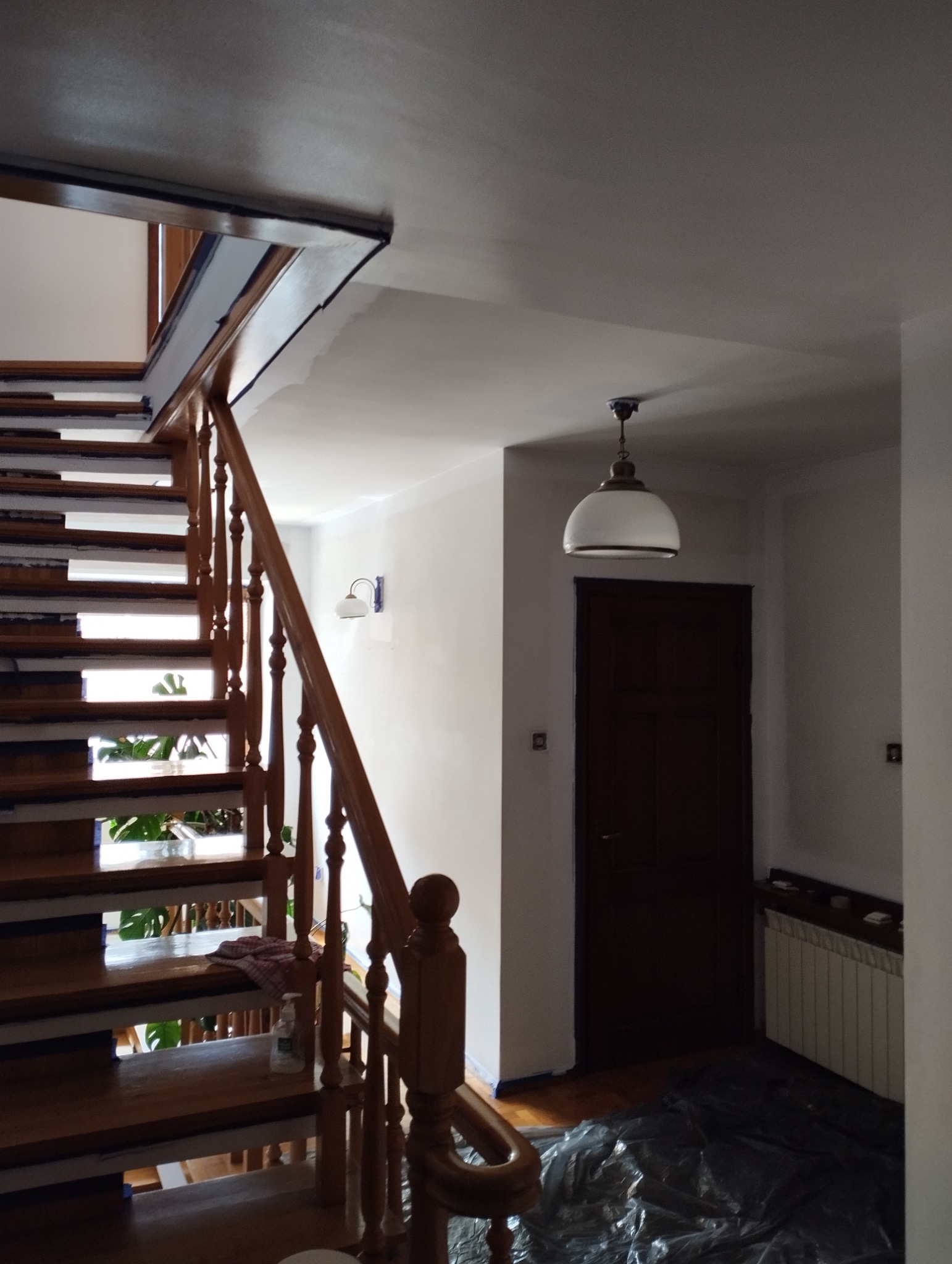 Klatka schodowa w trakcie malowania: drewniane schody zabezpieczone taśmą, świeżo pomalowane ściany na biało, widoczna lampa i folia ochronna na podłodze.