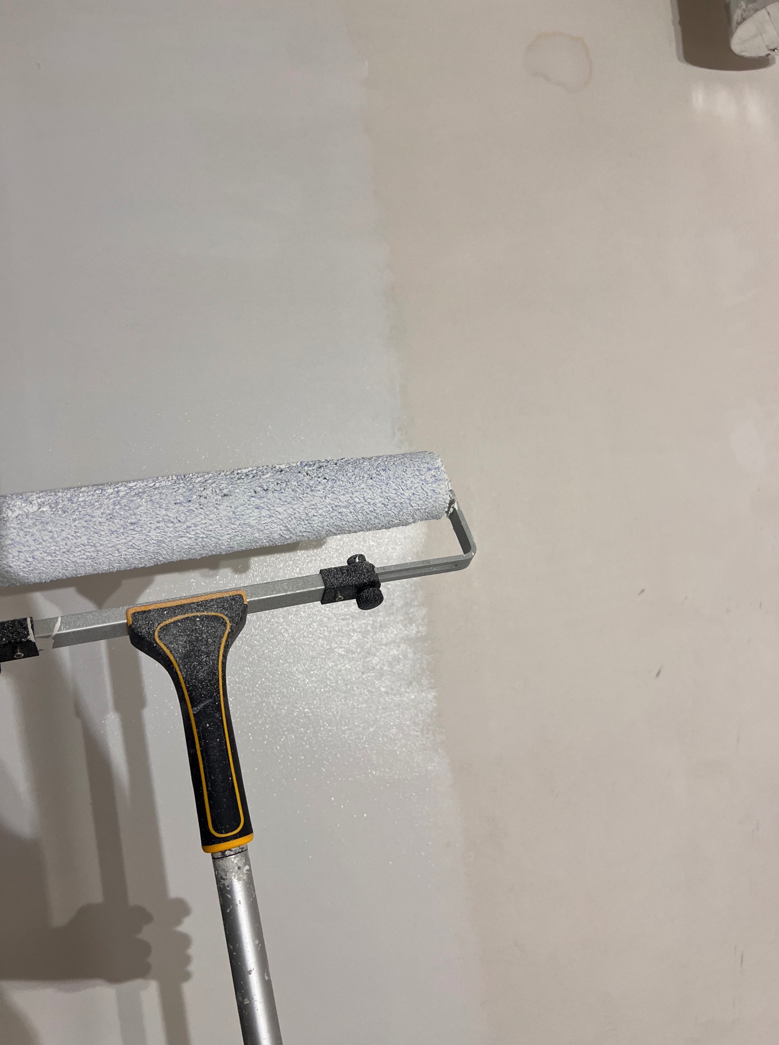 Wałek malarski z teleskopową rączką w trakcie malowania ściany na jasny odcień szarości, widoczna różnica w kolorze przed i po malowaniu.