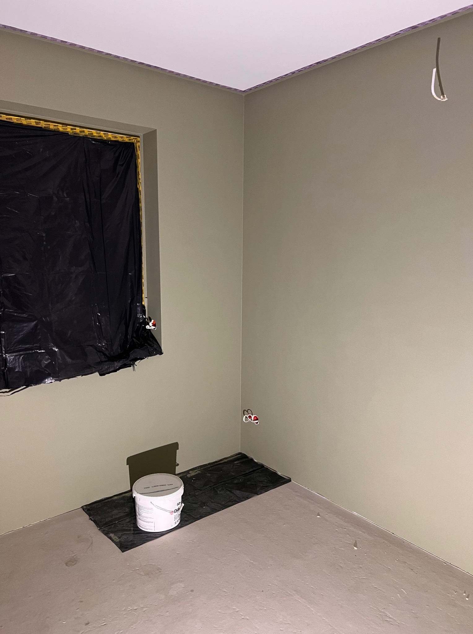 Pokój po malowaniu na oliwkowy odcień, okno zabezpieczone folią, wiadro farby na podłodze. Widoczne gniazdka elektryczne i wystający kabel.