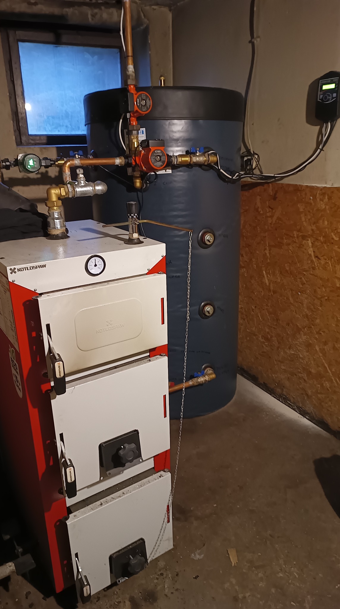 Kocioł na paliwo stałe marki Kotłospaw z podłączonym zasobnikiem ciepłej wody i osprzętem. Widoczne rury, pompy obiegowe i sterownik.