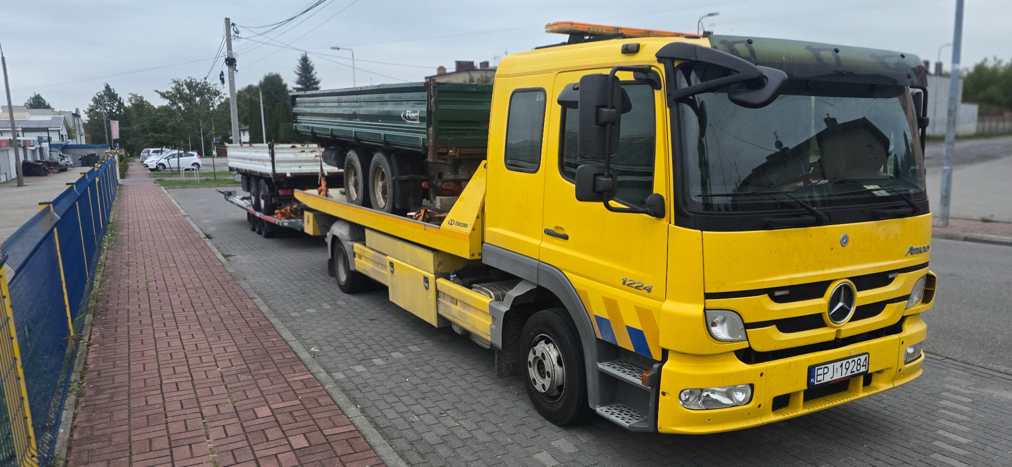 Żółta laweta Mercedes-Benz z platformą załadowaną przyczepą i zieloną skrzynią ładunkową, zaparkowana na ulicy z kostki brukowej. Widoczne tablice rejestracyjne.