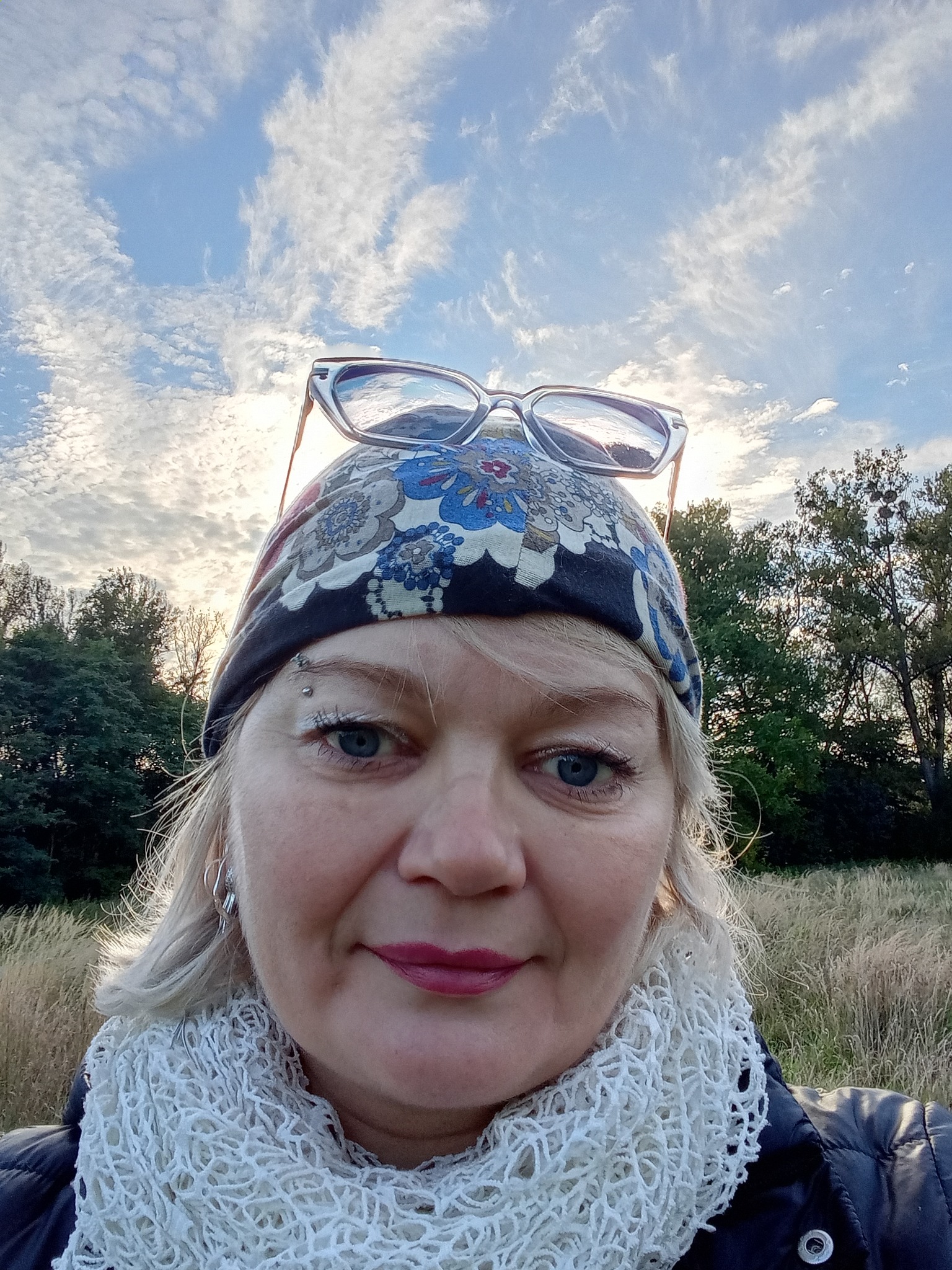 Portret kobiety z okularami na głowie, w chustce i szaliku, na tle łąki i nieba. Widoczne drzewa w oddali. Uśmiech, piercing brwi. Nietypowy kadr z dołu.