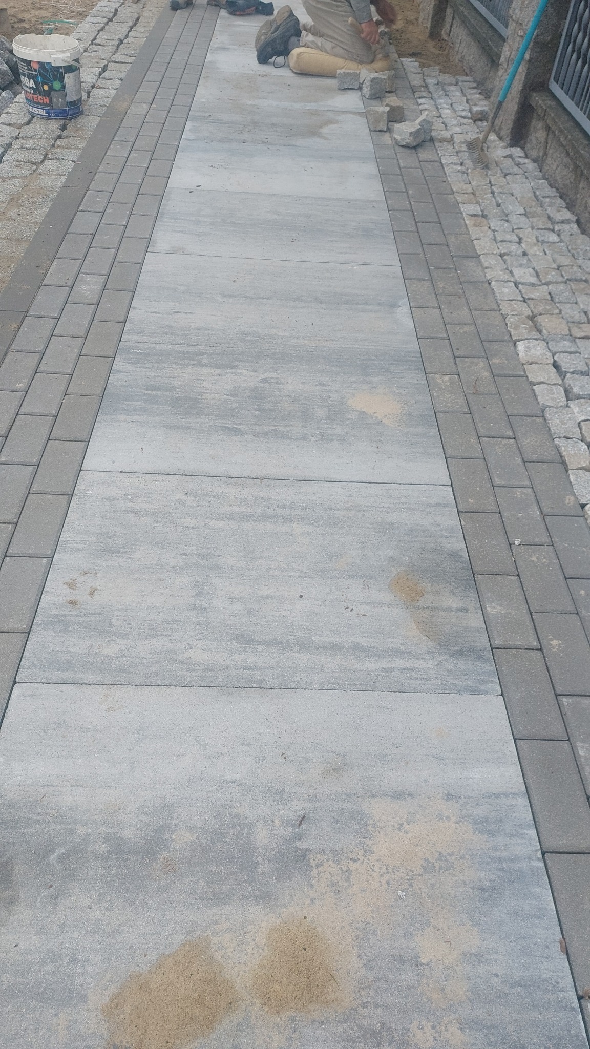 Ścieżka z dużych, szarych płyt betonowych obramowana kostką brukową i granitową. Widoczny etap prac z narzędziami i materiałami budowlanymi.