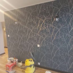 P.P.H.U. GLOB3. RAFAŁ GUGUŁA - Ściana z tapetą w motywy gałązek, oświetlona lampami. Na podłodze widoczny odkurzacz i akcesoria malarskie, co sugeruje prace wykończeniowe w pomieszczeniu.