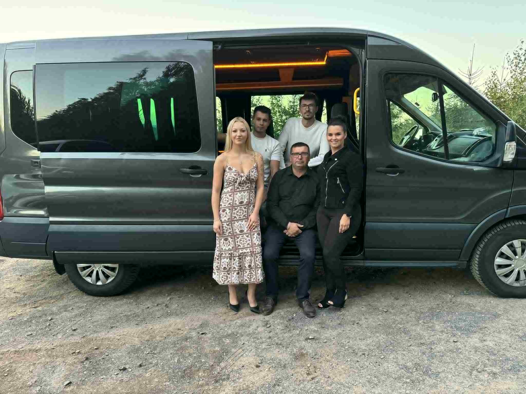 Grupa pięciu osób pozuje przy otwartym, szarym busie z podświetlanym wnętrzem. Widoczne drzewa w tle. Ujęcie z boku, na zewnątrz pojazdu.