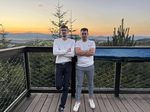 Dwóch mężczyzn na platformie widokowej z panoramą gór o zachodzie słońca. Informacyjna tablica z opisem krajobrazu w tle. Ubrani w codzienne stroje, pozują z założonymi rękami.