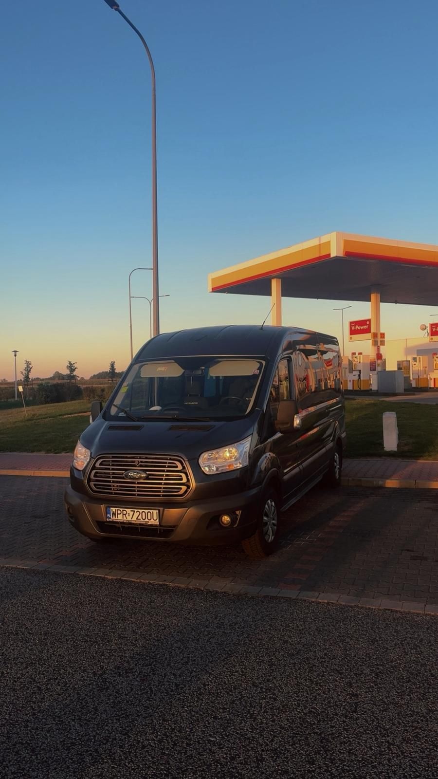 Czarny bus Ford Transit na tle stacji benzynowej Shell o zachodzie słońca. Widoczny numer rejestracyjny WPR 7200U. Ujęcie z dołu, podkreślające wielkość pojazdu.