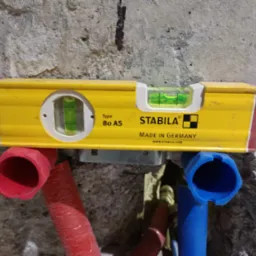 Żółta poziomica Stabila na tle surowej ściany, zamontowana nad czerwonymi i niebieskimi rurami instalacji wodnej. Widoczne oznaczenia producenta i poziomica w użyciu.