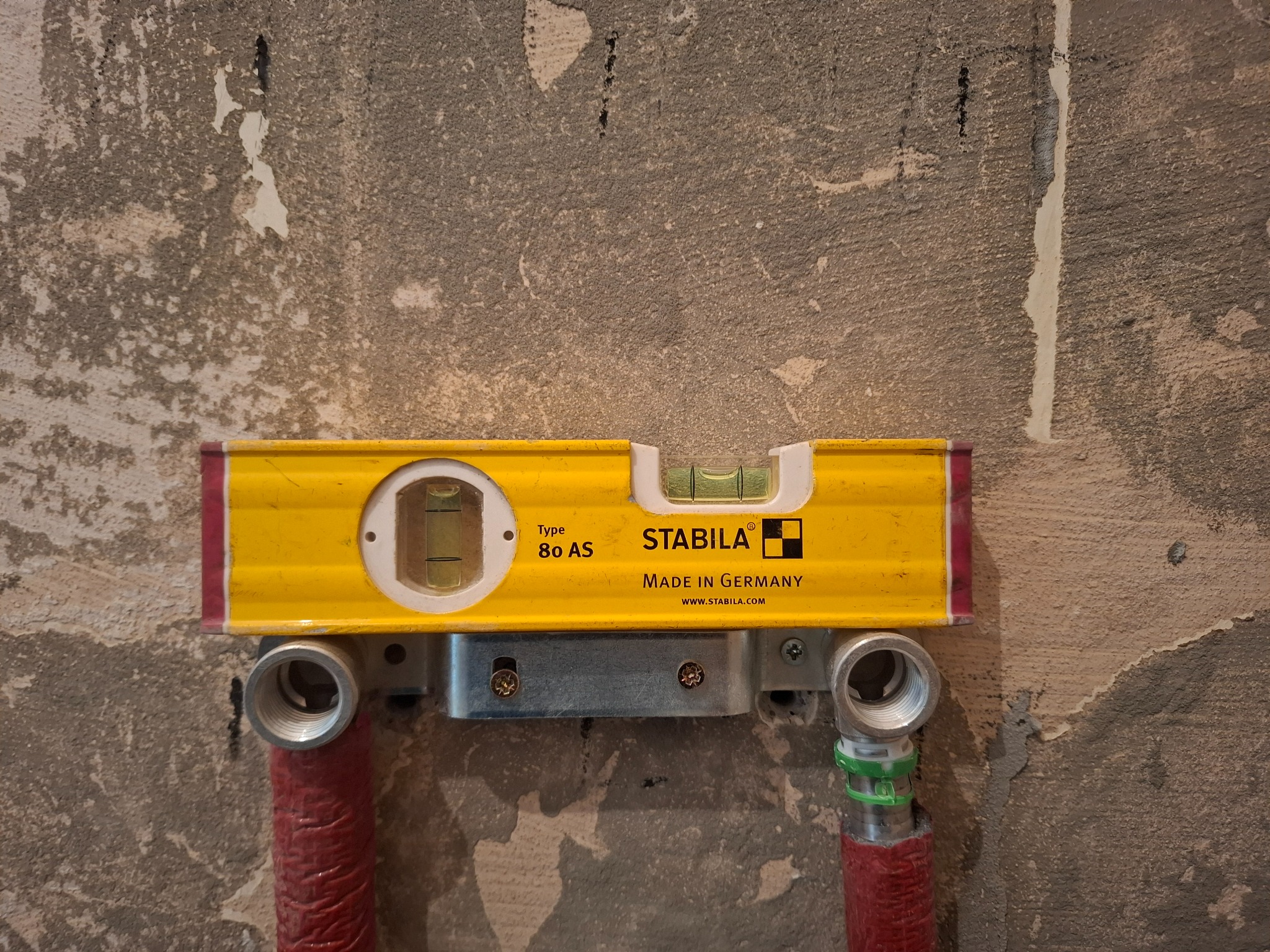 Poziomica Stabila na tle surowej ściany, zamontowana na rurach instalacji. Widoczne złącza i fragmenty rur w czerwonej izolacji. Precyzyjny montaż instalacji.