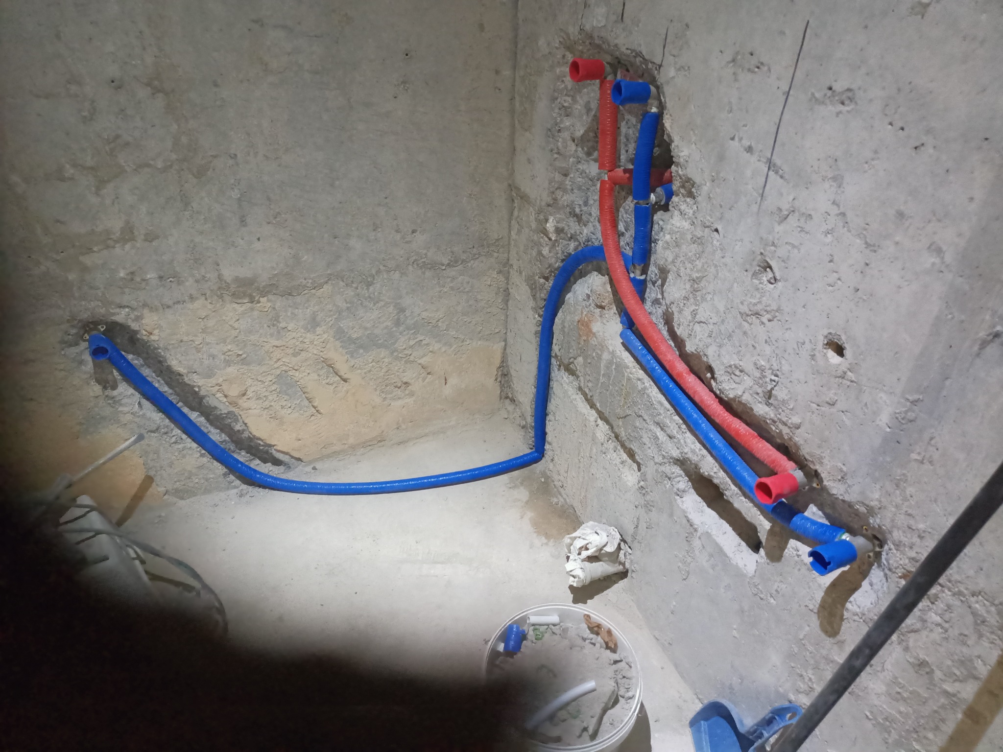 Instalacja wodna z rur PEX w surowym wnętrzu. Czerwone i niebieskie rury prowadzone wzdłuż ściany, widoczne narzędzia i akcesoria hydrauliczne. Surowy beton w tle.