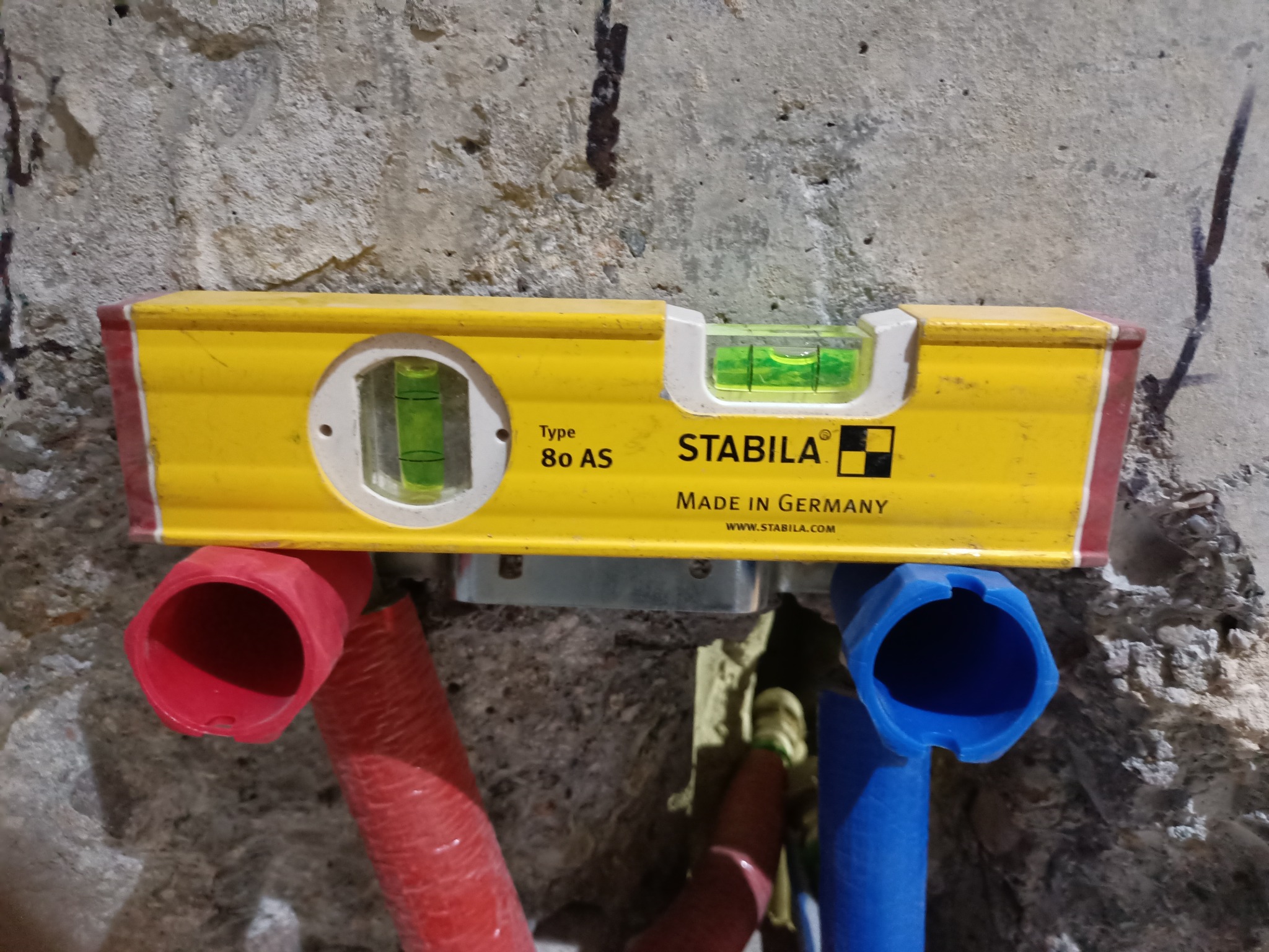 Żółta poziomica Stabila na tle surowej ściany, zamontowana nad czerwonymi i niebieskimi rurami instalacji wodnej. Widoczne oznaczenia producenta i poziomica w użyciu.