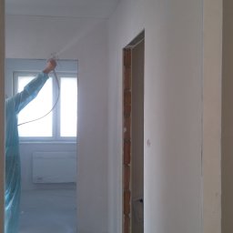 A-STAR Jan Szmolke - Prace wykończeniowe: malowanie natryskowe ścian wewnątrz pomieszczenia, widoczny pracownik w kombinezonie ochronnym i okno w tle. Ściany przygotowane do malowania.