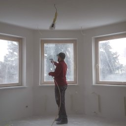 A-STAR Jan Szmolke - Malowanie natryskowe ścian w pokoju z trzema oknami, zabezpieczonymi folią. Widoczne kable oświetleniowe na suficie. Ściany świeżo pomalowane na biało.