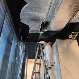 A-STAR Jan Szmolke - Pracownik na drabinie maluje natryskowo sufit na czarno. Sufit zabezpieczony folią, widoczne elementy instalacji. Ujęcie z dołu, podkreślające wysokość pomieszczenia.