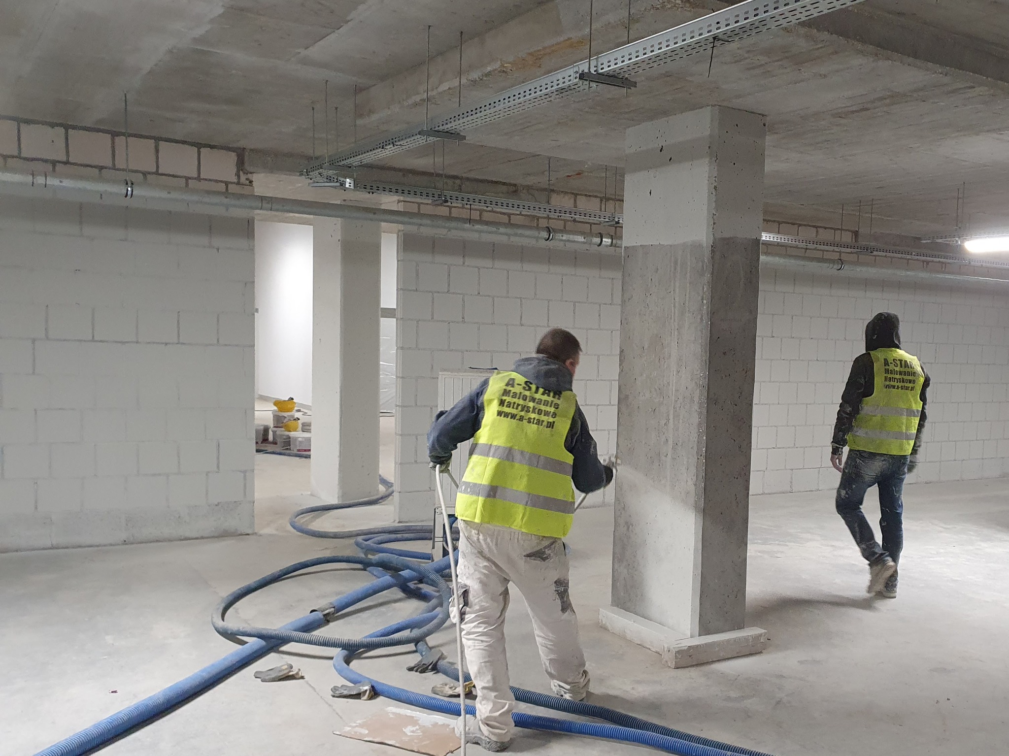 Pracownicy w kamizelkach malują natryskowo betonowy filar w surowym wnętrzu. Widoczne węże, ściany z białego bloczka i instalacje pod sufitem. Etap prac wykończeniowych.