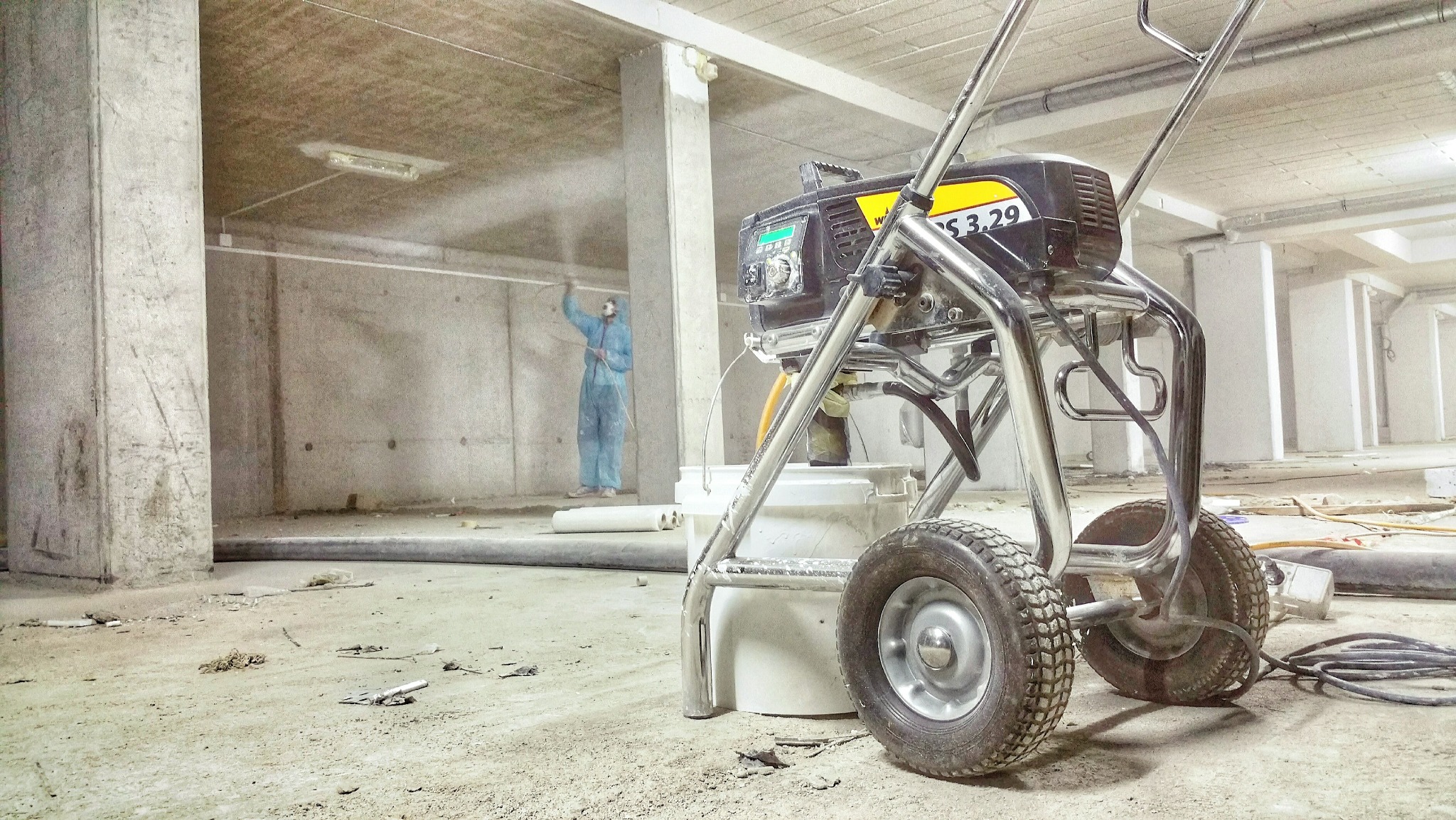 Malowanie natryskowe ścian w surowym wnętrzu. W tle pracownik w kombinezonie ochronnym. Na pierwszym planie agregat malarski z widocznym manometrem.