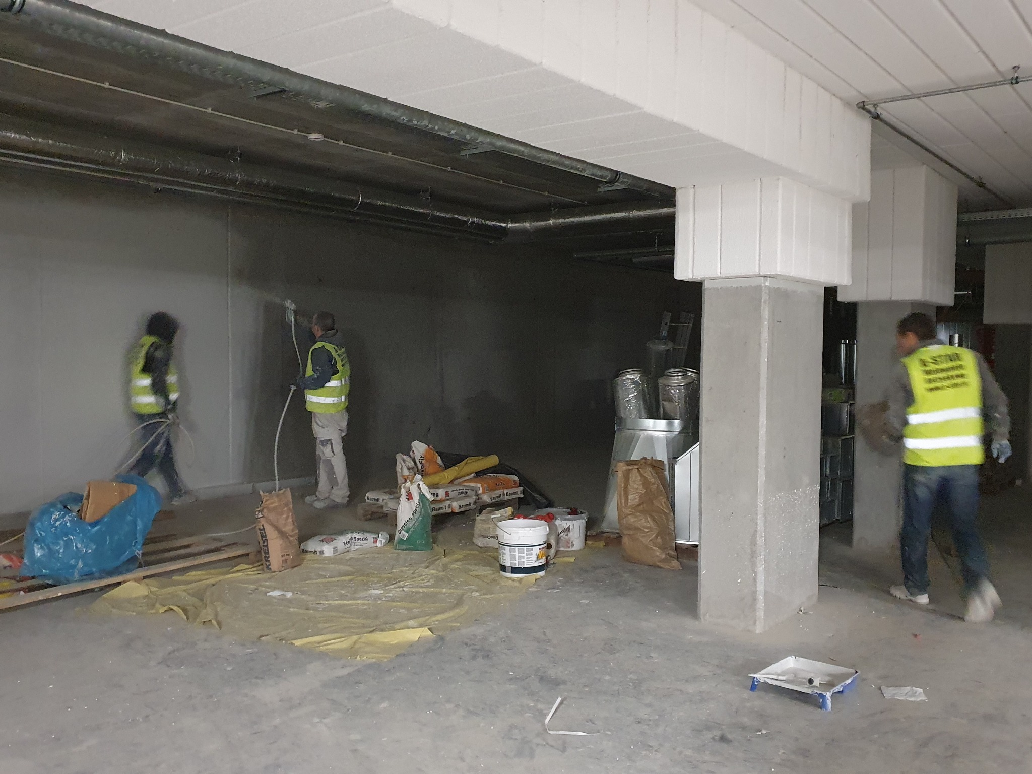 Prace wykończeniowe w hali: dwóch pracowników w kamizelkach nanosi gładź agregatem na ścianę. Widoczne worki z zaprawą, wiaderka i osłony.