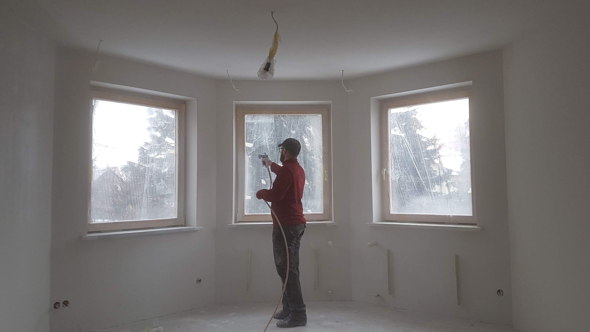 Malowanie natryskowe ścian w pokoju z trzema oknami, zabezpieczonymi folią. Widoczne kable oświetleniowe na suficie. Ściany świeżo pomalowane na biało.