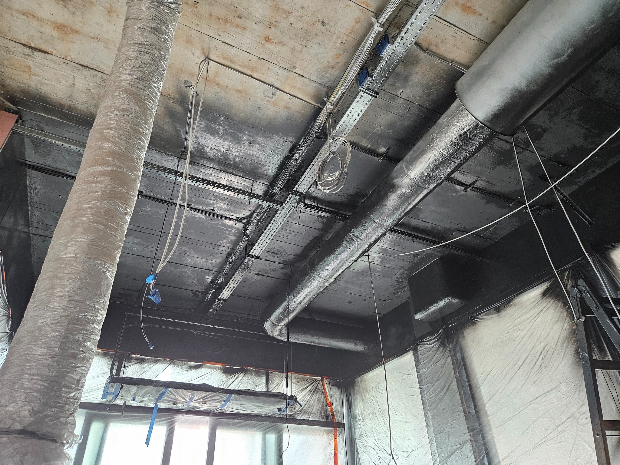Malowanie sufitu na czarno w pomieszczeniu z instalacjami wentylacyjnymi. Sufit drewniany, zabezpieczony folią. Widoczne kable i drabina. Ujęcie z dołu.