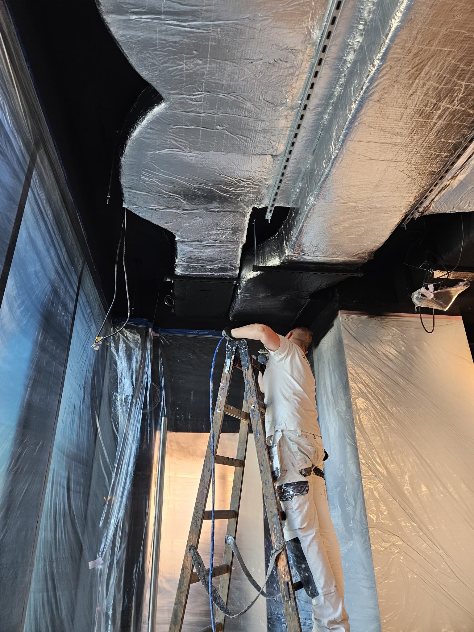 Pracownik na drabinie maluje natryskowo sufit na czarno. Sufit zabezpieczony folią, widoczne elementy instalacji. Ujęcie z dołu, podkreślające wysokość pomieszczenia.