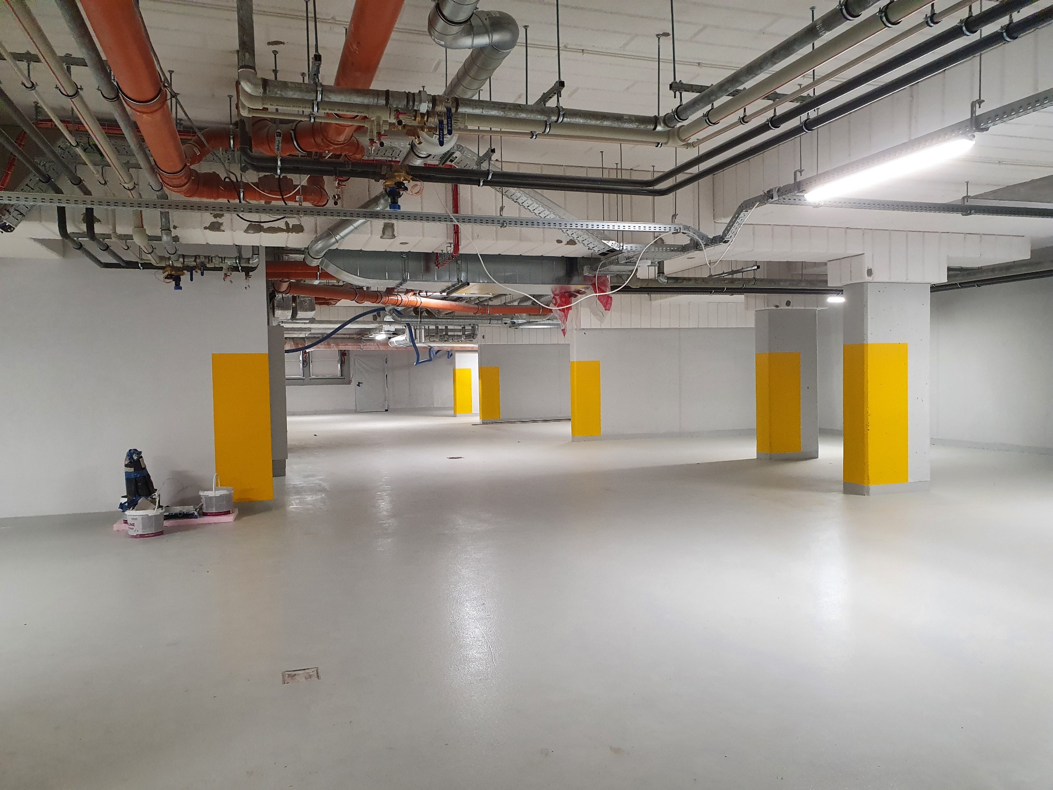 Jasne, przestronne wnętrze po malowaniu, z żółtymi akcentami na filarach. Widoczne instalacje pod sufitem i narzędzia malarskie w rogu. Minimalistyczny, industrialny styl.