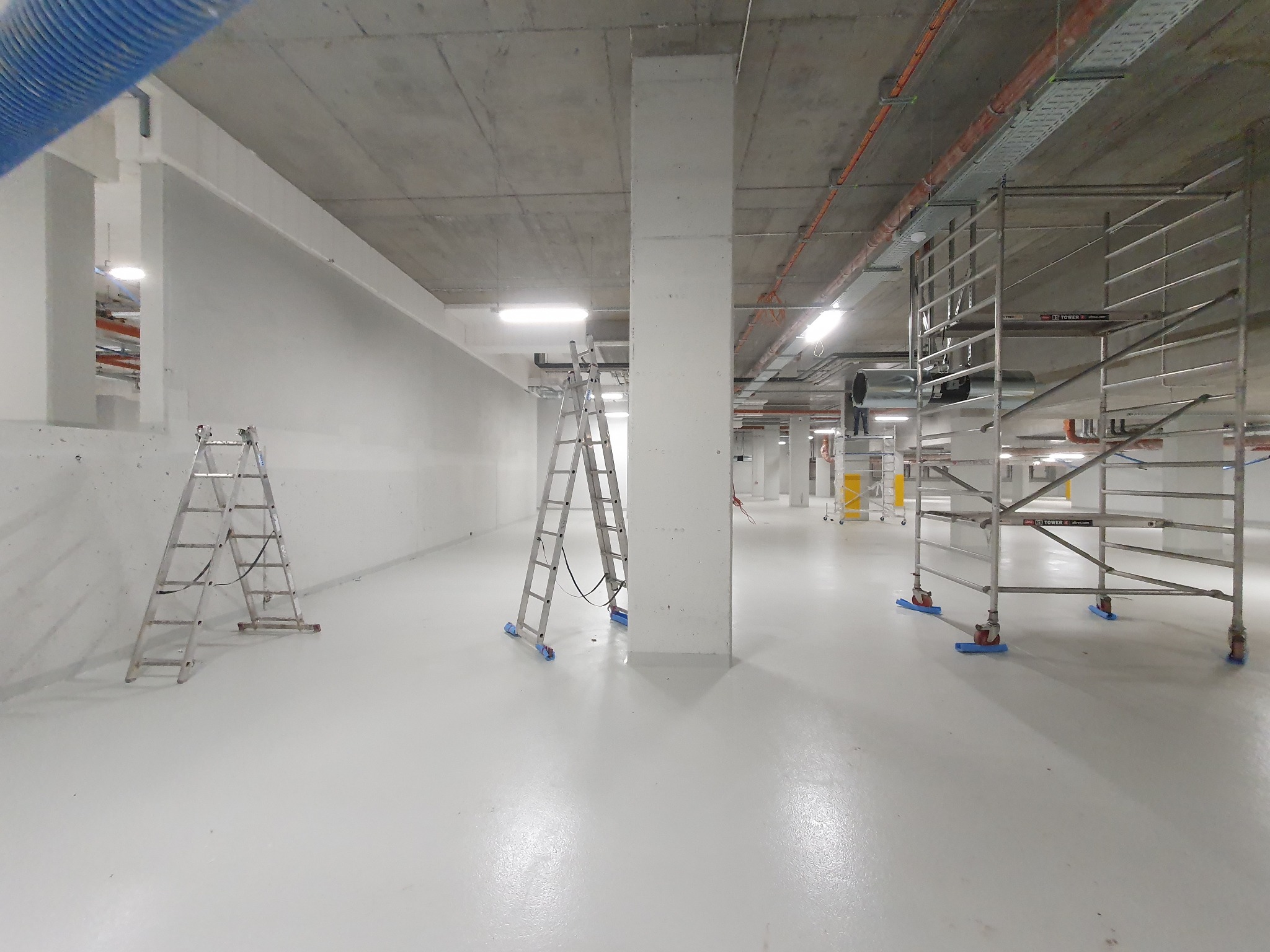 Białe, przestronne wnętrze w trakcie malowania: drabiny, rusztowania, gładkie ściany i podłoga. Widoczne instalacje pod sufitem. Minimalistyczny, industrialny charakter.