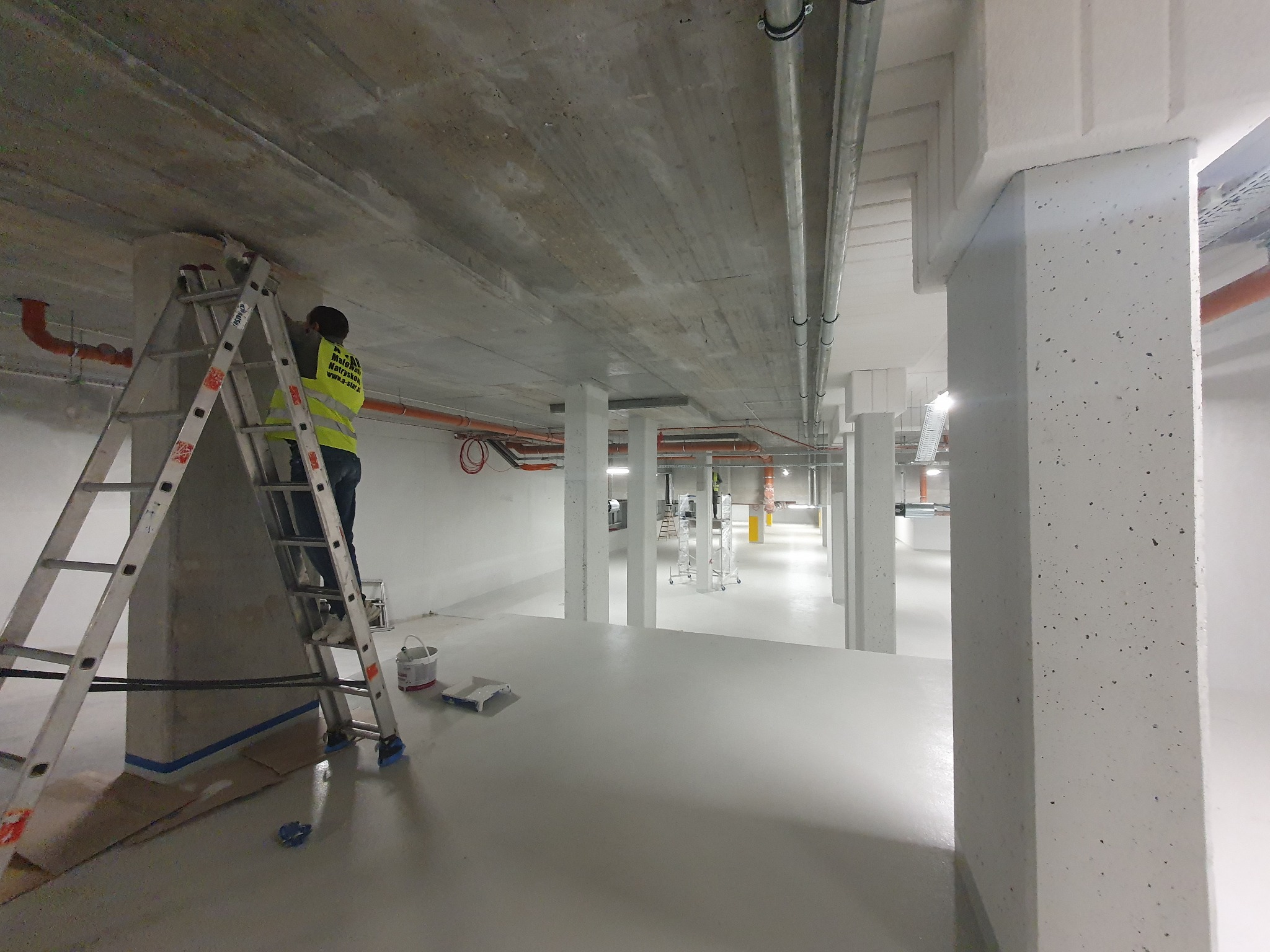 Malowanie kolumny w hali przez pracownika na drabinie. Widoczne zabezpieczenia podłoża, puszka z farbą i wałek. W tle rząd kolumn i instalacje pod sufitem.