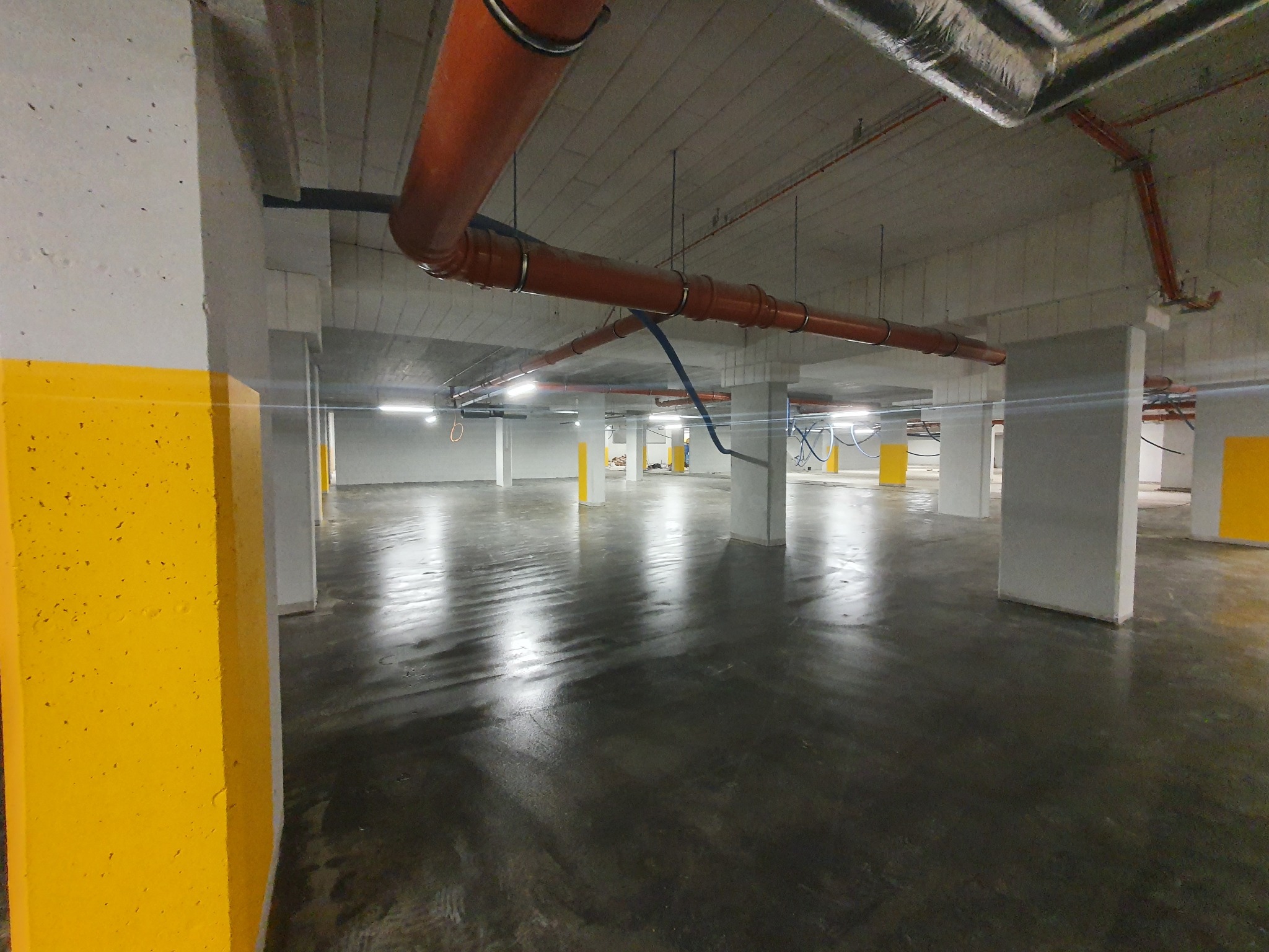 Wykończone, surowe wnętrze parkingu podziemnego z pomalowanymi na żółto filarami, widocznymi instalacjami pod sufitem i betonową posadzką.