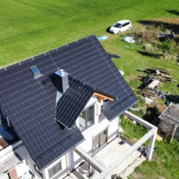 Nowoczesny dom z ciemną dachówką ceramiczną, widok z góry. Na dachu okno połaciowe i komin. W tle zielony trawnik i biały samochód. Częściowo widoczne rusztowania.