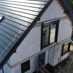 P.H.U Dach-System Sebastian Szaforz - Nowoczesny dom w trakcie budowy z ciemnoszarą dachówką i oknem dachowym. Widoczne białe ściany z betonu komórkowego i czarne ramy okienne. Ujęcie z góry.