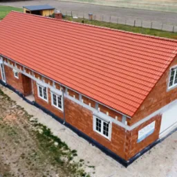 Budynek w trakcie budowy z czerwoną dachówką, widok z góry. Częściowo otynkowane ściany, okna wstawione, teren wokół budynku przygotowany. Widoczna tablica z nazwą firmy.