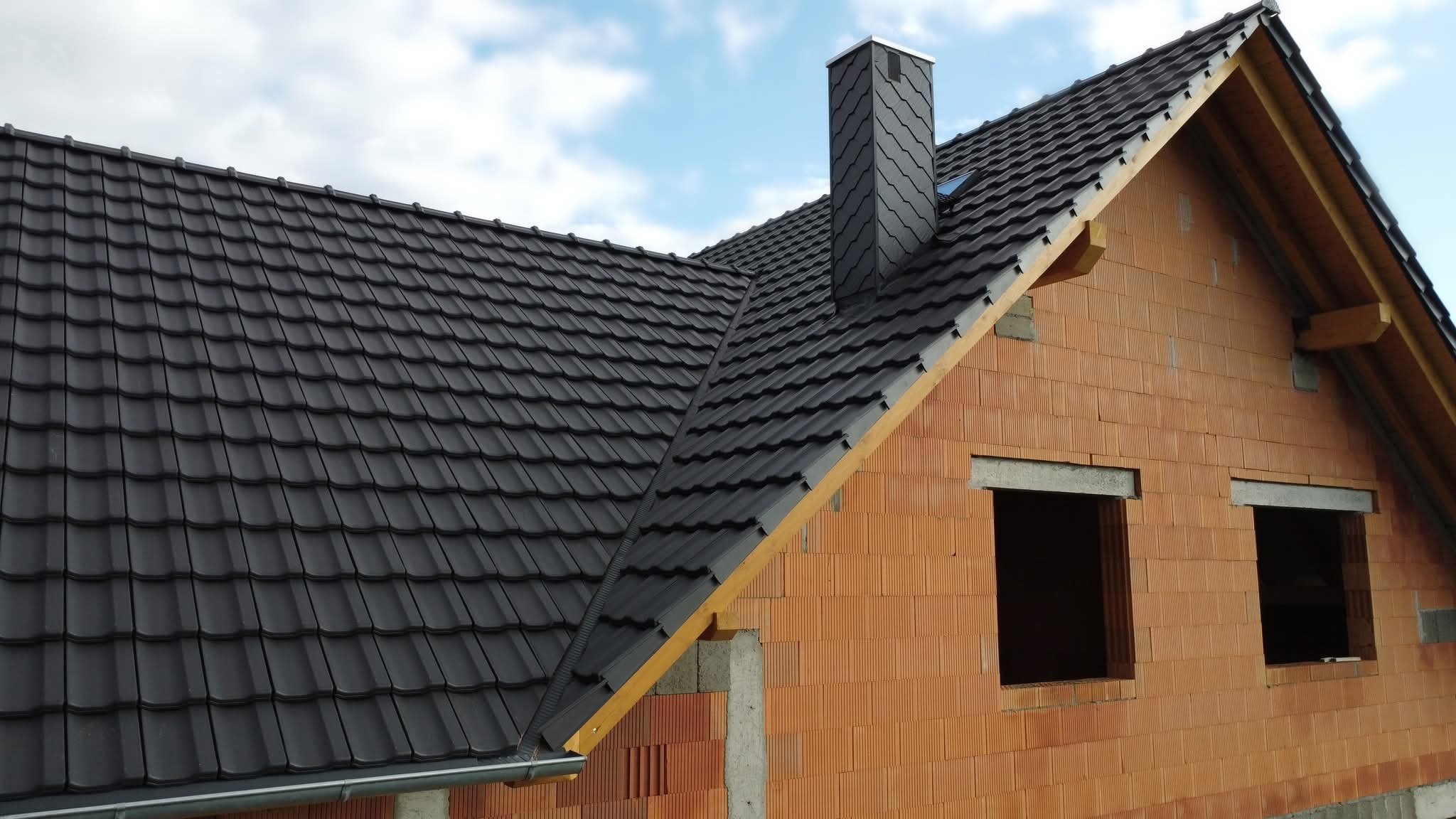Nowy, grafitowy dach z kominem na domu w trakcie budowy. Widoczne rynny i okno dachowe. Czerwona cegła dziurawka na ścianach. Błękitne niebo w tle.