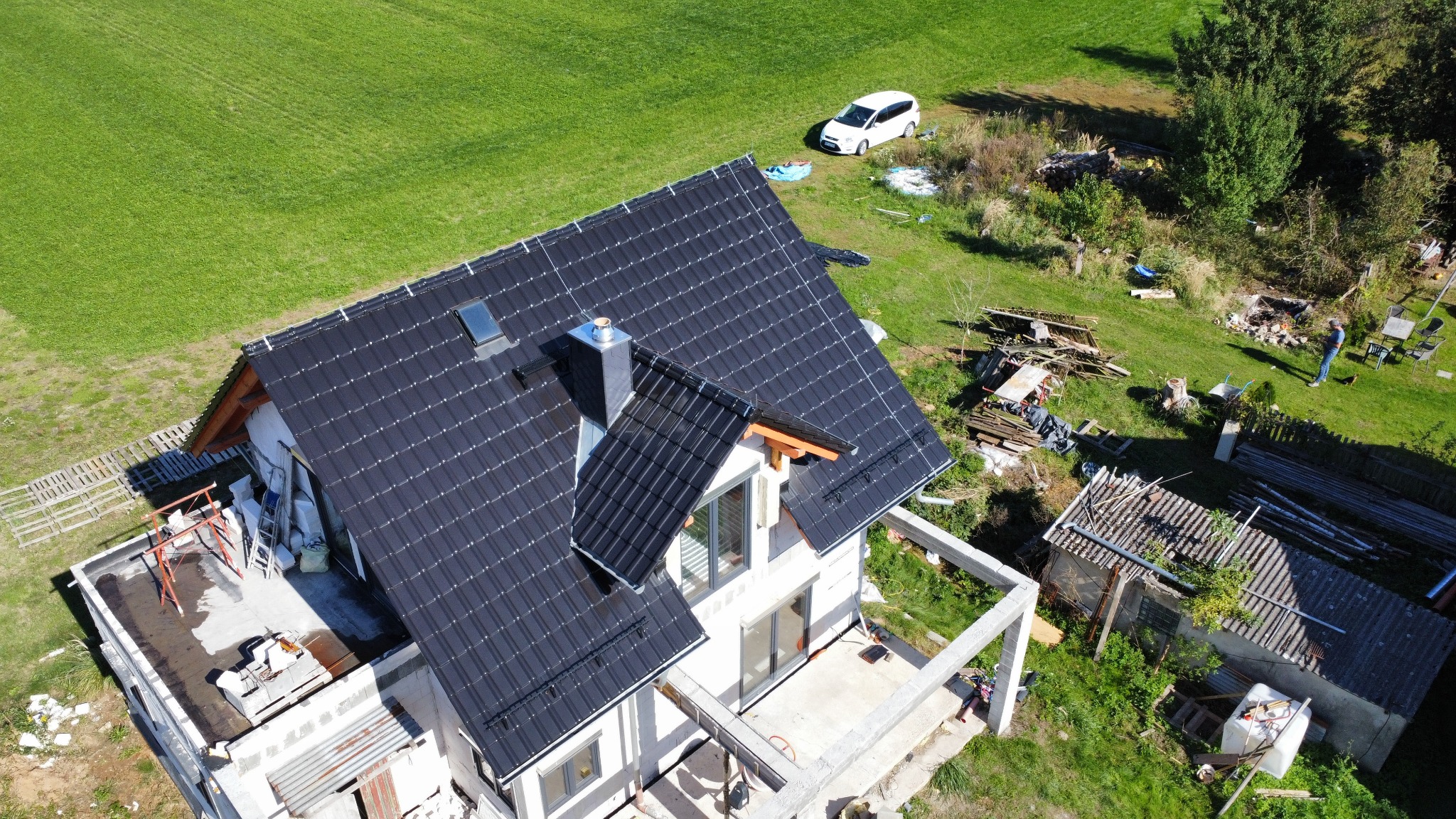 Nowoczesny dom z ciemną dachówką ceramiczną, widok z góry. Na dachu okno połaciowe i komin. W tle zielony trawnik i biały samochód. Częściowo widoczne rusztowania.
