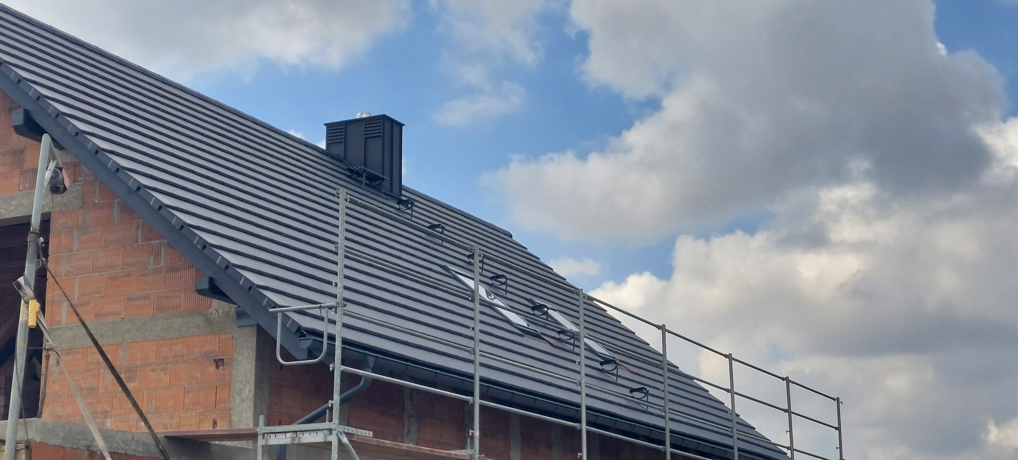 Ciemna dachówka ceramiczna na nowym dachu z kominem, widok z rusztowania na tle błękitnego nieba z chmurami. Częściowo widoczna ściana budynku w trakcie budowy.