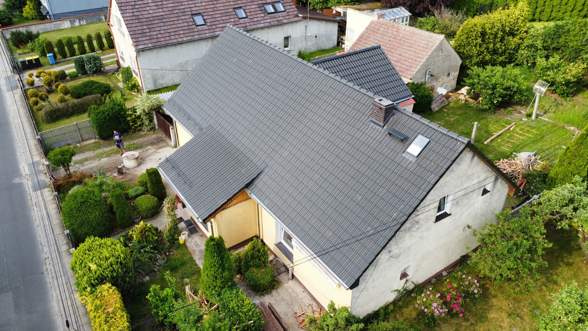 Dom jednorodzinny z szarym dachem z dachówki ceramicznej, widok z góry. Na dachu okno dachowe i komin. W tle zieleń ogrodu i inne budynki.